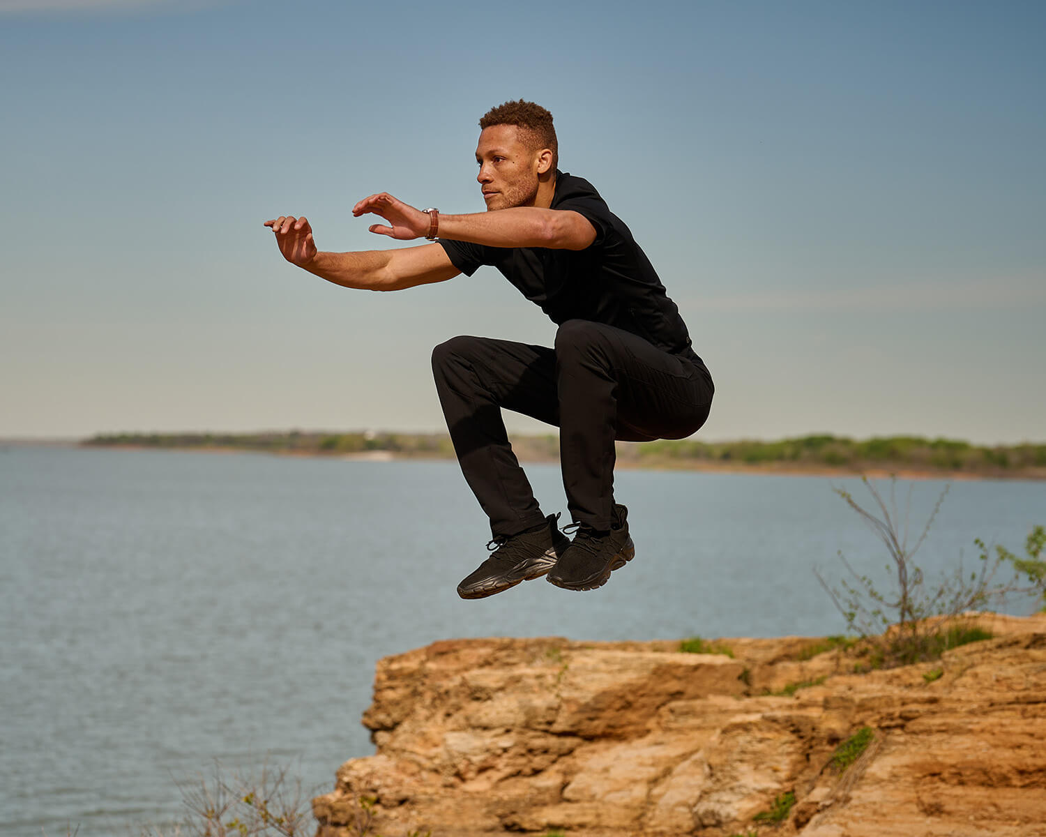 Man in all black Black Crest Joggers and black short sleeve shirt in a jump squat
